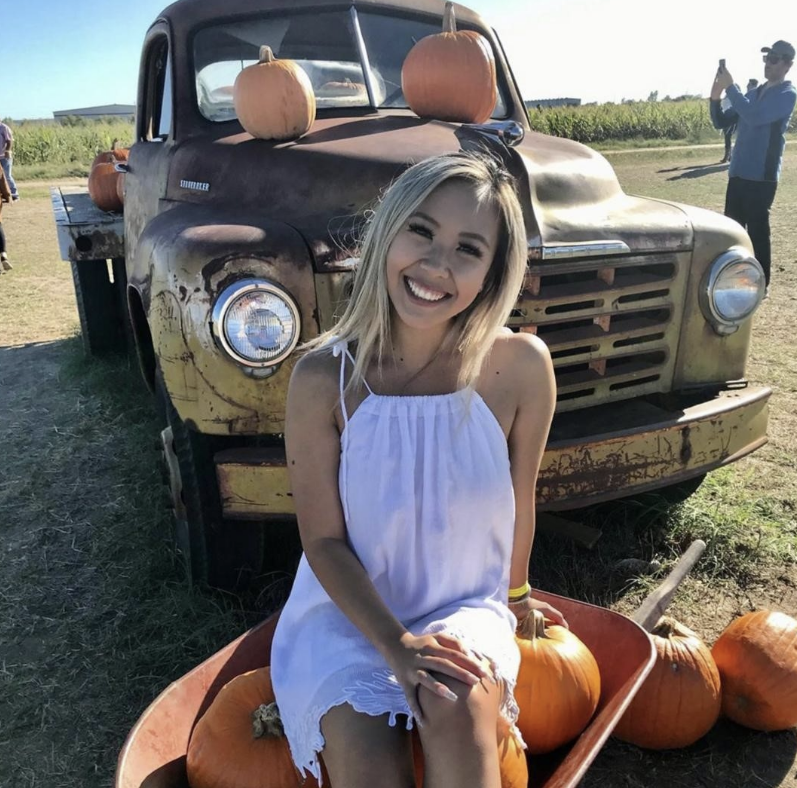 A smiling woman sitting by pumpkins with a classic truck behind her